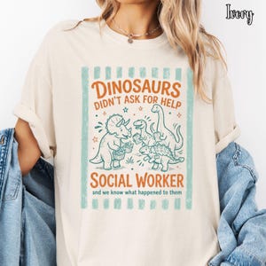 May include: An ivory t-shirt featuring a graphic print of four dinosaurs with the text "DINOSAURS DIDN'T ASK FOR HELP SOCIAL WORKER and we know what happened to them" in orange and teal, framed by vertical teal stripes on a light-colored background.