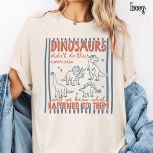 May include: An ivory t-shirt featuring a graphic print with blue vertical stripes and illustrations of three dinosaurs. The text reads: DINOSAURS didn't do their exercises and we know what HAPPENED TO THEM. The word Ivory is printed in the top right corner.