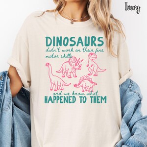 May include: An ivory t-shirt featuring a graphic print with four pink dinosaur illustrations. The text reads: DINOSAURS didn't work on their fine motor skills and we know what HAPPENED TO THEM. The shirt is layered under a blue denim jacket.