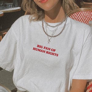 May include: White t-shirt with the red text "BIG FAN OF HUMAN RIGHTS". The person is wearing silver necklaces and sunglasses with a white frame. The person is sitting in a red and white chair at a cafe.