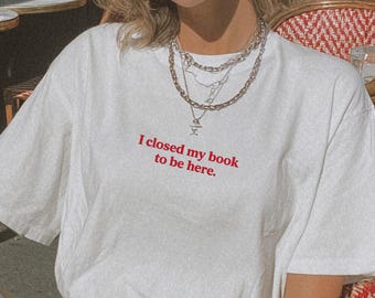 May include: White t-shirt with the red text "I closed my book to be here." The person is wearing silver-rimmed sunglasses and layered necklaces. The person is sitting at a cafe table with a glass of water.