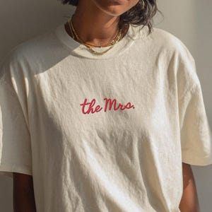 Puede incluir: Camiseta color crema con el texto bordado "the Mrs." en escritura roja. La camiseta tiene mangas cortas y un corte relajado. La modelo lleva joyas doradas y pantalones oscuros.
