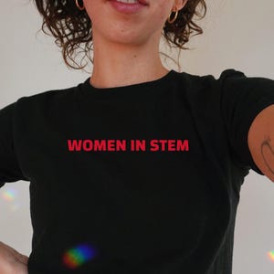 May include: Black crew neck t-shirt with the red text "WOMEN IN STEM". The person wearing the shirt is also wearing blue jeans.