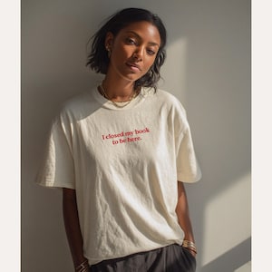 May include: Cream-colored t-shirt with the text "I closed my book to be here" in red. The shirt has a relaxed fit and is worn by a person. The person is wearing gold jewelry and dark pants.