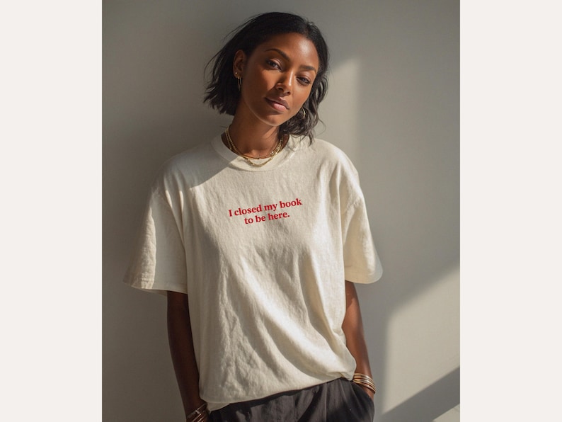 May include: Cream-colored t-shirt with the text "I closed my book to be here" in red. The shirt has a relaxed fit and is worn by a person. The person is wearing gold jewelry and dark pants.