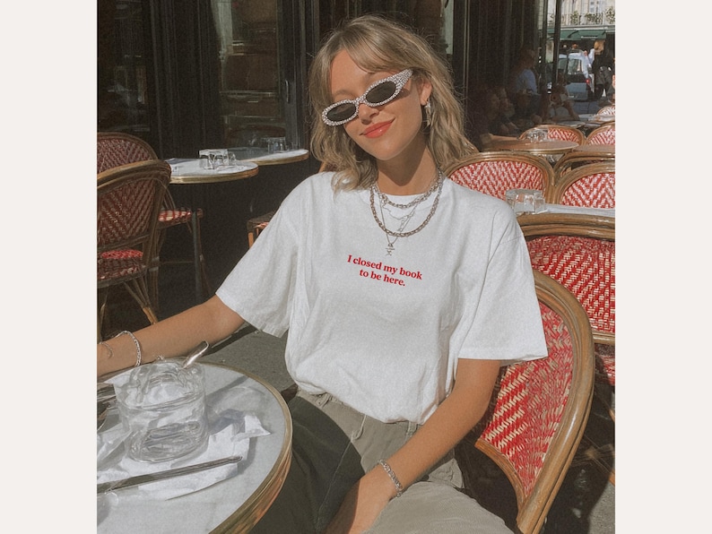May include: White t-shirt with the red text "I closed my book to be here." The person is wearing silver-rimmed sunglasses and layered necklaces. The person is sitting at a cafe table with a glass of water.