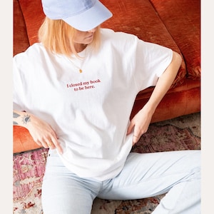 May include: White t-shirt with the red text "I closed my book to be here." The shirt is worn with light blue jeans and a light blue baseball cap. A gold necklace is visible.