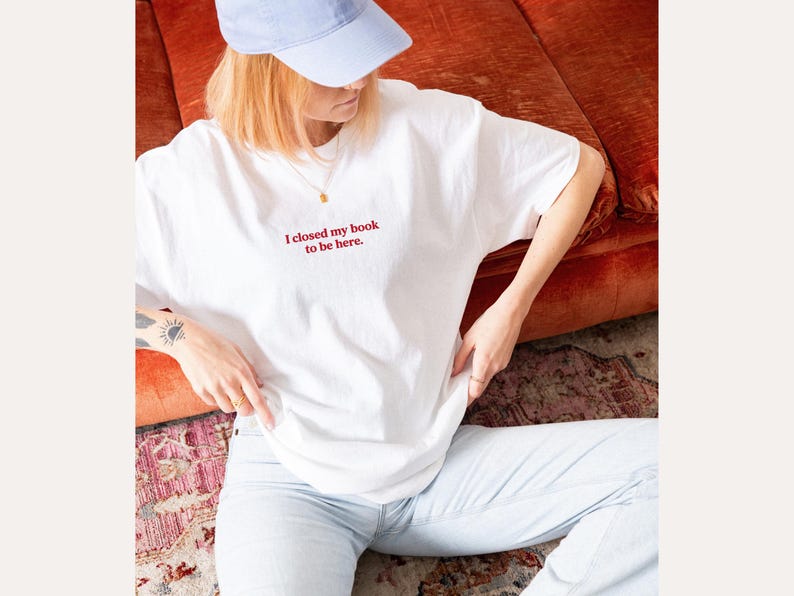 May include: White t-shirt with the red text "I closed my book to be here." The shirt is worn with light blue jeans and a light blue baseball cap. A gold necklace is visible.