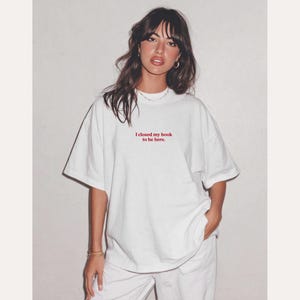 May include: White oversized t-shirt with the red text "I closed my book to be here." The shirt is paired with white pants. The model is wearing gold hoop earrings and a necklace.