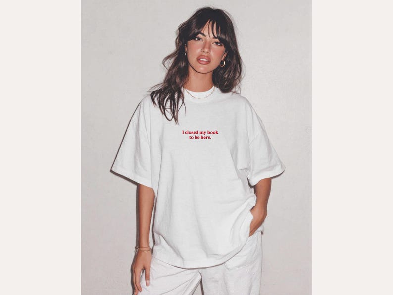 May include: White oversized t-shirt with the red text "I closed my book to be here." The shirt is paired with white pants. The model is wearing gold hoop earrings and a necklace.