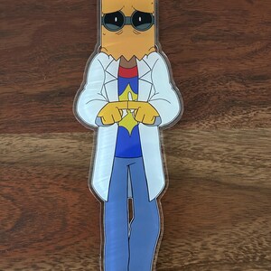 K&ouml;nnte beinhalten: Ein klarer Acryl-Anh&auml;nger mit einer Cartoon-Figur mit Papiert&uuml;tenkopf, Sonnenbrille und wei&szlig;em Laborkittel. Die Figur tr&auml;gt ein blaues Hemd, eine blaue Hose und rote Schuhe. Oben ist ein roter Verschluss angebracht.
