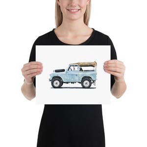 May include: A watercolour print of a vintage Land Rover in light blue. The off-road vehicle features a tan canvas roof, black tyres, and a spare tyre on the bonnet. The artwork is presented on a white background.