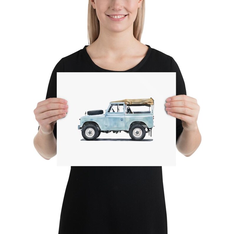 May include: A watercolour print of a vintage Land Rover in light blue. The off-road vehicle features a tan canvas roof, black tyres, and a spare tyre on the bonnet. The artwork is presented on a white background.