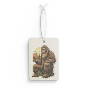 May include: Rectangular air freshener with a cartoon Bigfoot holding a pint of beer. The Bigfoot is brown and seated in front of a forest scene with a yellow sun. The air freshener has a white string for hanging.