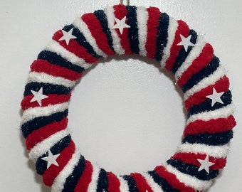 May include: A red, white and blue yarn wreath with white stars. The wreath is made of a soft, fluffy yarn and is perfect for a patriotic display.
