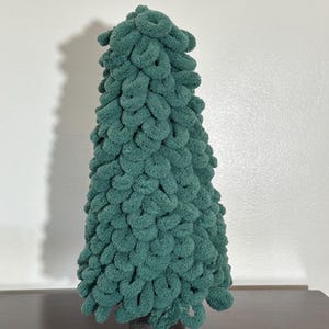以下が含まれることがあります： 茶色の土台に置かれた緑色のニット製のクリスマスツリー。ツリーは柔らかくふわふわした糸でできており、独特の質感があります。