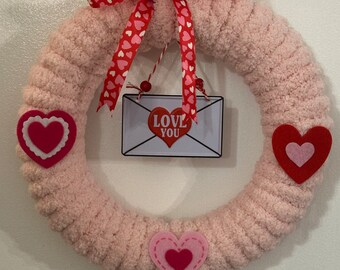 May include: Pink fluffy wreath with a red and white ribbon and a sign that says "Love You" with a red heart. The wreath is decorated with red and pink felt hearts.