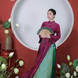 Op de afbeelding: Een traditionele Vietnamese jurk voor dames, met een lange, vloeiende bordeauxrode top en een turquoise en groene rok met patroon. De jurk wordt gecombineerd met een waaier en een zilveren ketting. De achtergrond omvat een grote witte cirkel en bloemstukken.