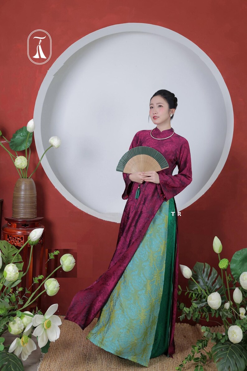Op de afbeelding: Een traditionele Vietnamese jurk voor dames, met een lange, vloeiende bordeauxrode top en een turquoise en groene rok met patroon. De jurk wordt gecombineerd met een waaier en een zilveren ketting. De achtergrond omvat een grote witte cirkel en bloemstukken.