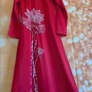 May include: A long, red dress with long sleeves and a high neckline, adorned with white floral embroidery and silver embellishments. The dress is hanging on a hanger against a gold background.