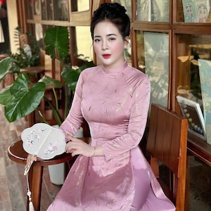 May include: A woman wearing a long-sleeved, pink dress with a high neckline and gold accents. She is seated, holding a white fan with floral designs. The dress has a high slit and a matching pink underskirt.