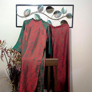 May include: Two-piece red and green traditional Vietnamese Áo dài. The long-sleeved top and matching pants feature a floral pattern. The Áo dài is hanging on a wooden chair in front of a decorative wall hanging.