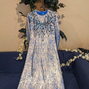 May include: A long, silver and blue dress with long sleeves. The dress features a round neckline and is embellished with black and blue floral embroidery around the neckline. The dress is hanging on a hanger.