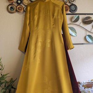 May include: A long, mustard-yellow dress with long sleeves and a mandarin collar. The fabric features a subtle floral pattern. A burgundy panel is visible on the side. The dress is hanging on a hanger.
