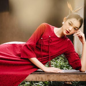 May include: A red dress with a floral pattern and three-quarter sleeves. The dress has a round neckline and decorative ties. The model is leaning on a wooden bench. The background is blurred, with greenery and a warm light.
