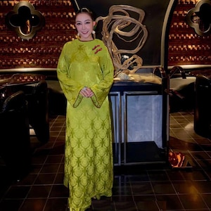 May include: A long, flowing, lime green dress with a floral pattern. The dress has long sleeves with flared cuffs and red trim. Two red floral embellishments are on the chest. The dress is worn by a person.