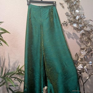 May include: A pair of high-waisted, wide-leg trousers in a deep emerald green color with a subtle iridescent sheen. The pants feature a structured waistband, a center back zipper closure, and a flowing, floor-length silhouette made of fabric.
