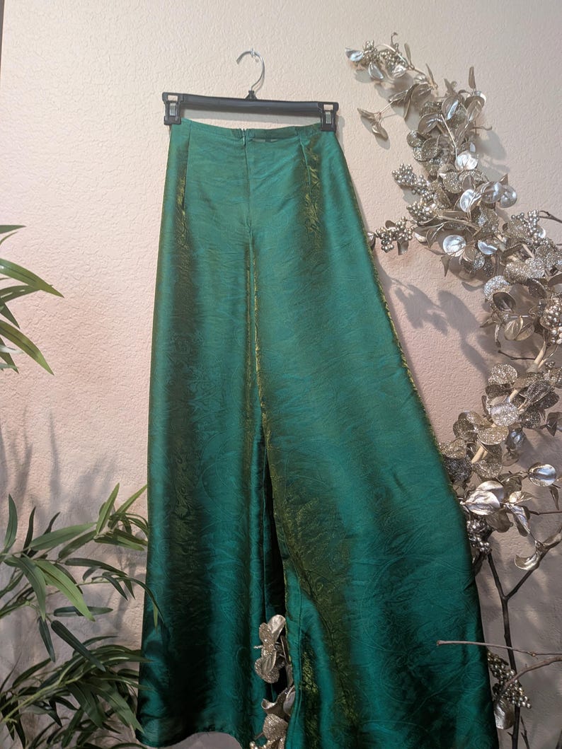 May include: A pair of high-waisted, wide-leg trousers in a deep emerald green color with a subtle iridescent sheen. The pants feature a structured waistband, a center back zipper closure, and a flowing, floor-length silhouette made of fabric.