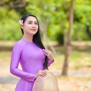 May include: A woman in a long-sleeved, lavender dress holds a woven conical hat. She has long black hair, a flower in her hair, and is looking upwards. The background is a blurred view of trees and foliage.