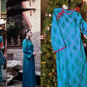 May include: A teal-colored, long-sleeved dress with a floral pattern and red trim. The dress features a mandarin collar and decorative frog closures. The dress is displayed on a hanger in front of a Christmas tree.