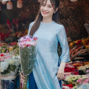 May include: A light blue tunic dress with a floral pattern, paired with dark blue pants. The dress has three-quarter sleeves and a high neckline. The person is holding a bouquet of pink and white flowers. The image includes the text "HUYEN THAI".