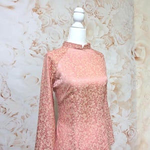 May include: A pink, long-sleeved dress with a mandarin collar and floral pattern. The dress is displayed on a white mannequin. The background features a floral wallpaper.