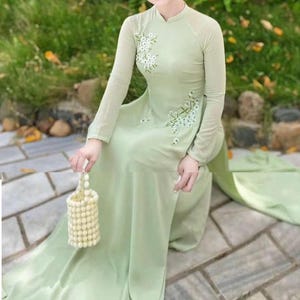 May include: A light green, long-sleeved dress with white floral embroidery. The dress features a high collar and a flowing skirt. A small, beaded handbag is held in the hand. The dress is suitable for formal occasions.