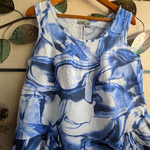 May include: A sleeveless top featuring a white and blue marbled pattern with swirling abstract shapes. The garment has a round neckline and a tiered, ruffled hemline at the bottom. A small tag is attached to the inner collar area of the shirt.