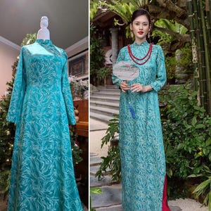 May include: A teal-colored, long-sleeved dress with a high collar and a flowing silhouette. The dress features a white, swirling pattern. The dress is displayed on a mannequin and worn by a person. The dress has a slit on the side.