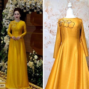 May include: A long, mustard yellow dress with long sleeves and a floral design on the chest. The dress is made of a shiny fabric and is shown on a mannequin and a person.