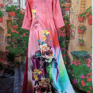 May include: A long, flowing pink dress with a floral print. The dress features a round neckline and long sleeves. The floral design includes large roses and other flowers in shades of yellow, purple, and white. The dress is displayed on a mannequin.