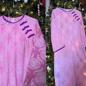 May include: A pink, long-sleeved dress with a mandarin collar and decorative frog closures. The dress features a subtle pattern and is trimmed with purple accents. The dress is displayed on a hanger.