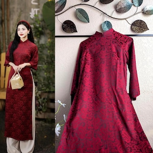 May include: A burgundy-colored, long-sleeved dress with a floral pattern. The dress has a mandarin collar and a slit up the side. The model is wearing a matching headband and carrying a small, gold-colored purse. The dress is paired with cream-colored pants.