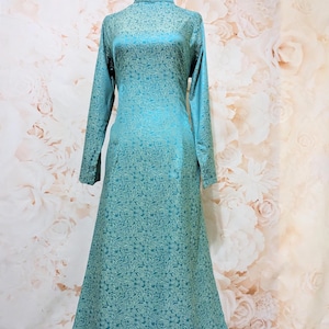 May include: A teal-colored, long-sleeved dress with a high neckline and a floral pattern. The dress is displayed on a mannequin against a floral background. The dress has a flowing, floor-length skirt.