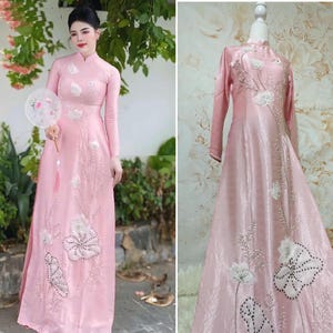 May include: A long, light pink dress with long sleeves and a mandarin collar. The dress features white floral embroidery and embellishments. The dress is shown on a mannequin and also worn by a person holding a fan.