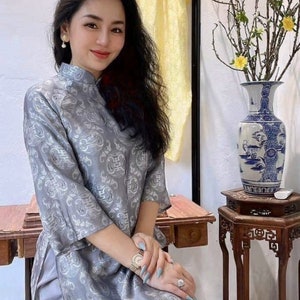 May include: A woman wearing a gray patterned dress with a mandarin collar. She is seated near a wooden table and a blue and white vase with branches and yellow flowers. A yellow garment hangs in the background.