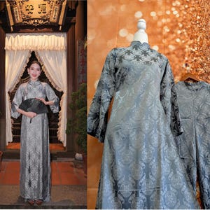 May include: A silver-gray, long-sleeved dress with a high collar and ornate pattern. The dress is displayed on a mannequin and also worn by a person holding a black fan. The dress has a matching jacket. The image is set in a traditional Asian setting.