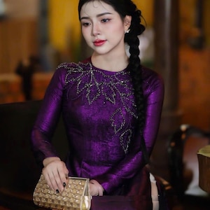 May include: A woman in a long-sleeved, deep purple dress with intricate silver embellishments around the neckline. She holds a gold, beaded clutch. Her dark hair is styled in a braid.