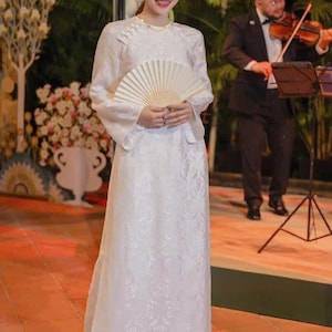 May include: A woman in a white, long-sleeved dress with a floral pattern, holding a folded fan. A violinist in a black suit plays in the background. The setting appears to be an outdoor event.
