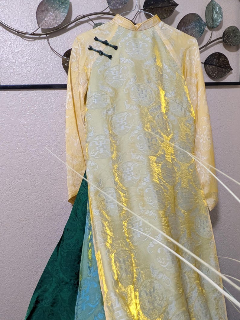 Op de afbeelding: Een lange, gele jurk met een mandarijnkraag en lange mouwen. De jurk heeft een subtiel, herhalend patroon en zwarte kikkerknopen. De rok is gelaagd met groene en blauwe stof, wat een contrast in kleur en textuur cre&euml;ert. De jurk hangt aan een hanger.